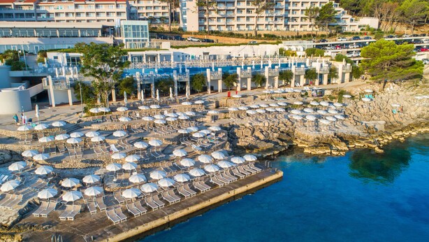 A coastal resort with multiple sun loungers and umbrellas, a rocky pier, and a backdrop of multi-story buildings. - Apartments Punta (Mali Losinj)
