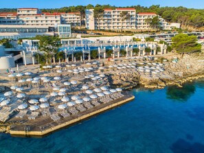 A coastal resort with multiple sun loungers and umbrellas, a rocky pier, and a backdrop of multi-story buildings. - Apartments Punta (Mali Losinj)