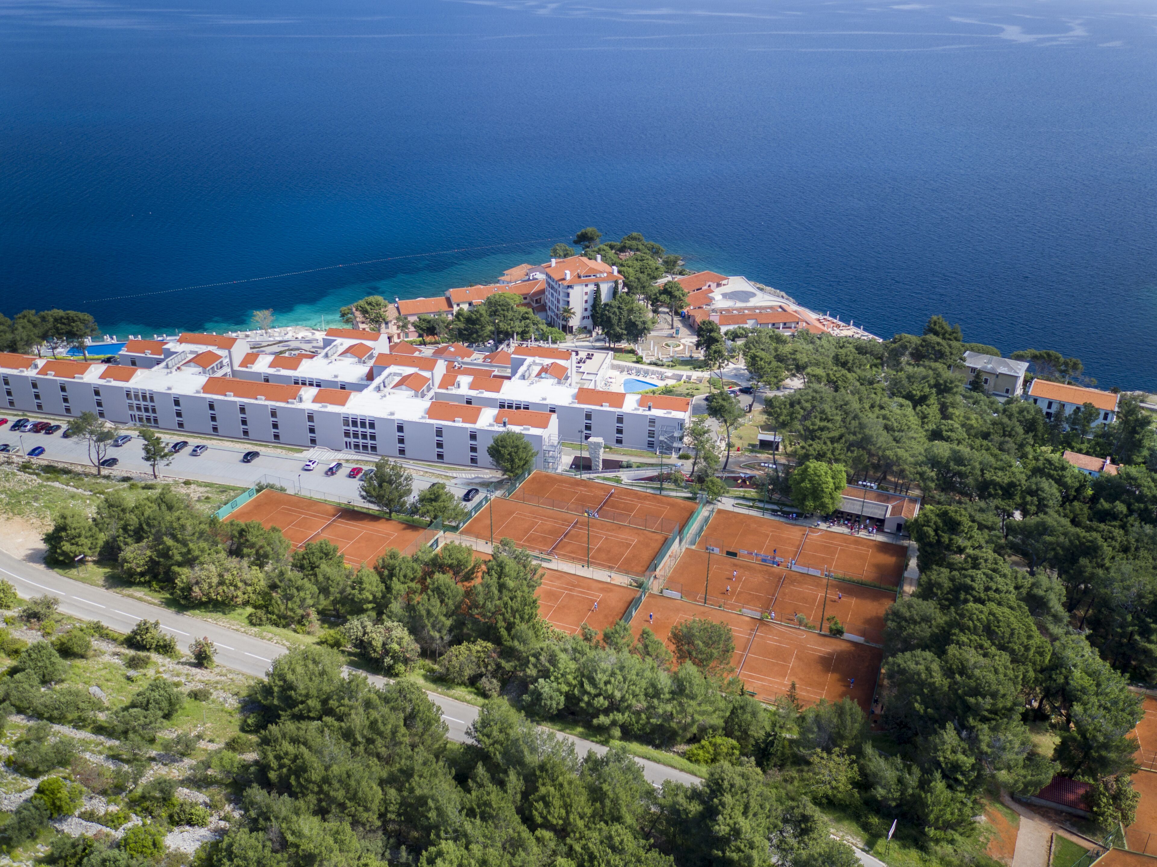 Ovenudblik over et kysthotel med flere bygninger, tennisbaner og en parkeringsplads.