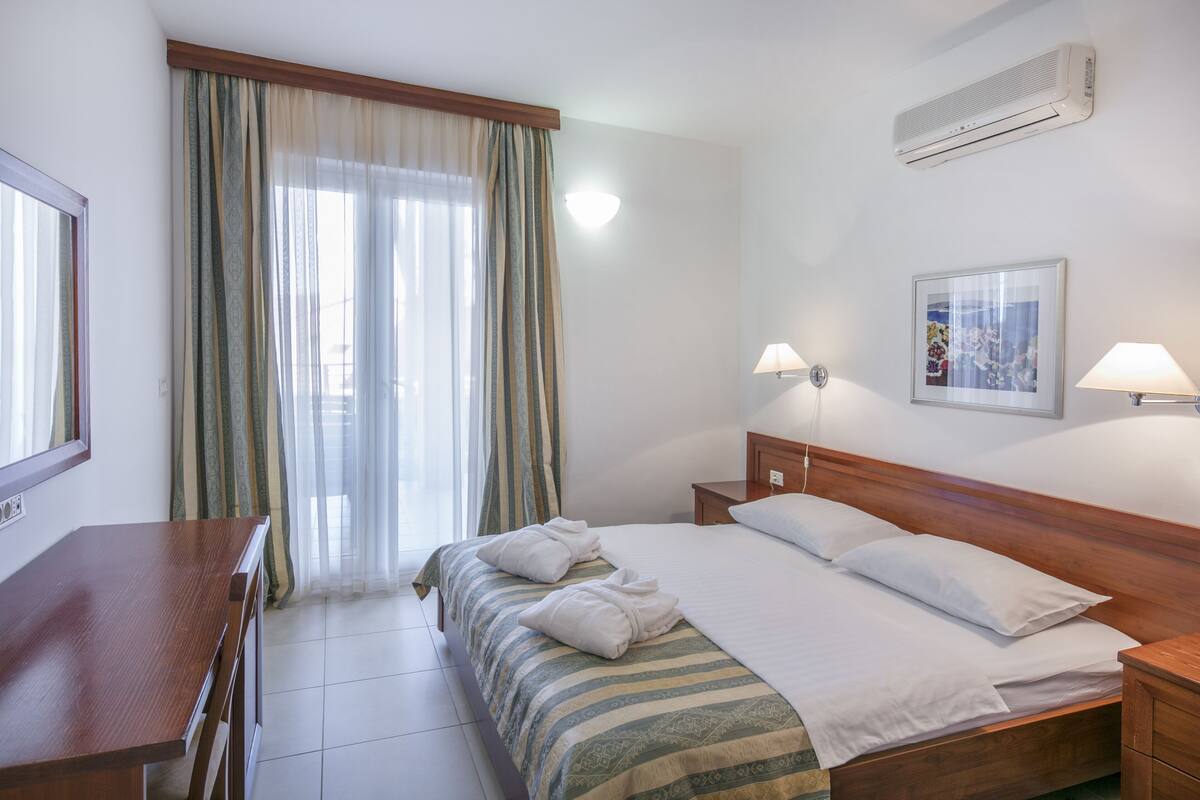A hotel room with a bed, bedside lamps, a wooden nightstand, a desk, a window with curtains, an air conditioner, and a framed picture on the wall.