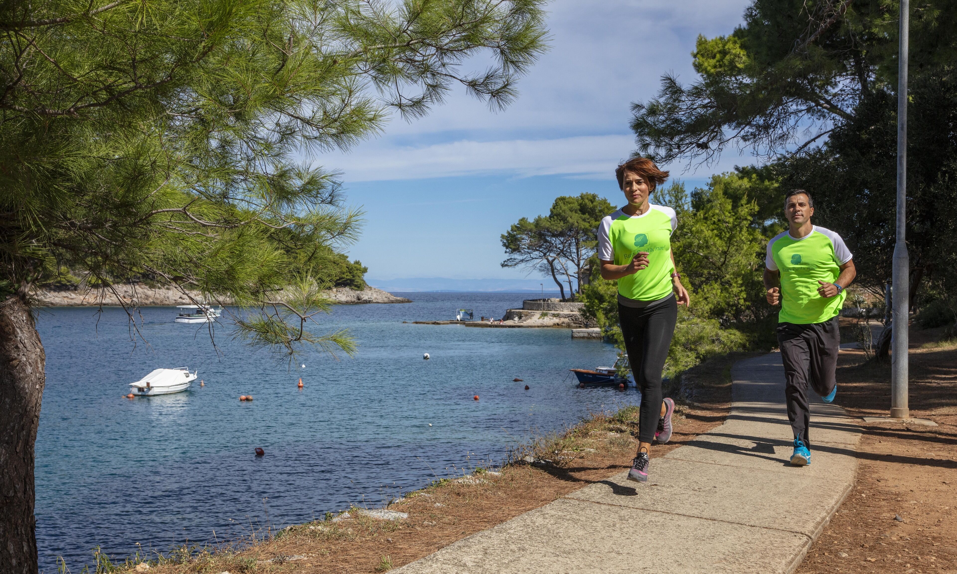 Δύο άτομα τρέχουν σε ένα παραλιακό μονοπάτι, με μια έκταση νερού και βάρκες να διακρίνονται στο βάθος.