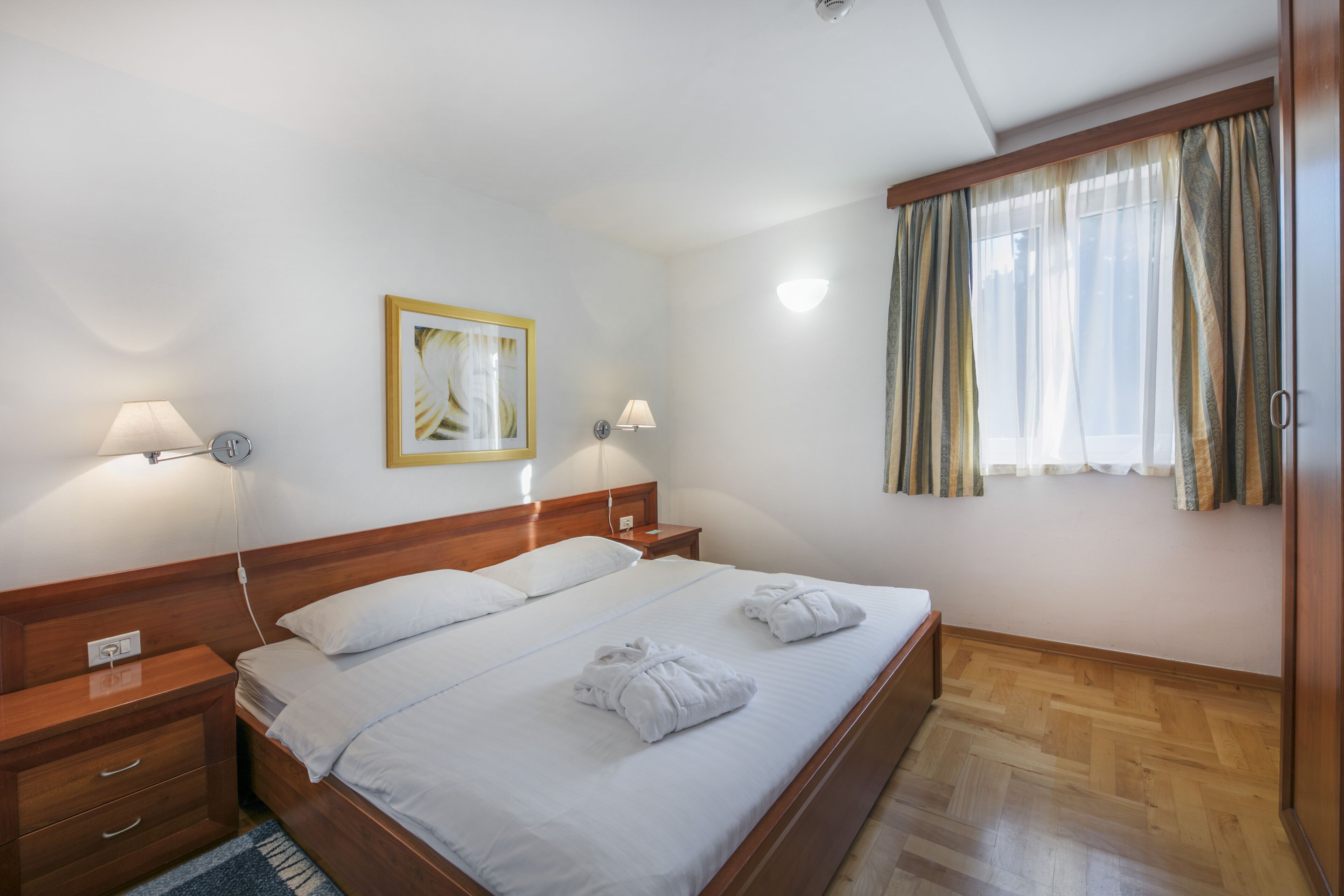 Une chambre d’hôtel avec un lit en bois, deux tables de chevet avec des lampes, un tableau encadré au mur et une fenêtre avec des rideaux.