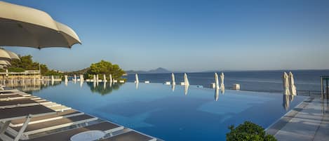 An infinity pool with lounge chairs and umbrellas, overlooking the sea and a mountain.