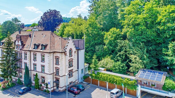 Un edificio de varios pisos con un estilo arquitectónico tradicional, que presenta tejado de tejas y fachada de piedra. Hay un área de estacionamiento al frente con varios automóviles estacionados.