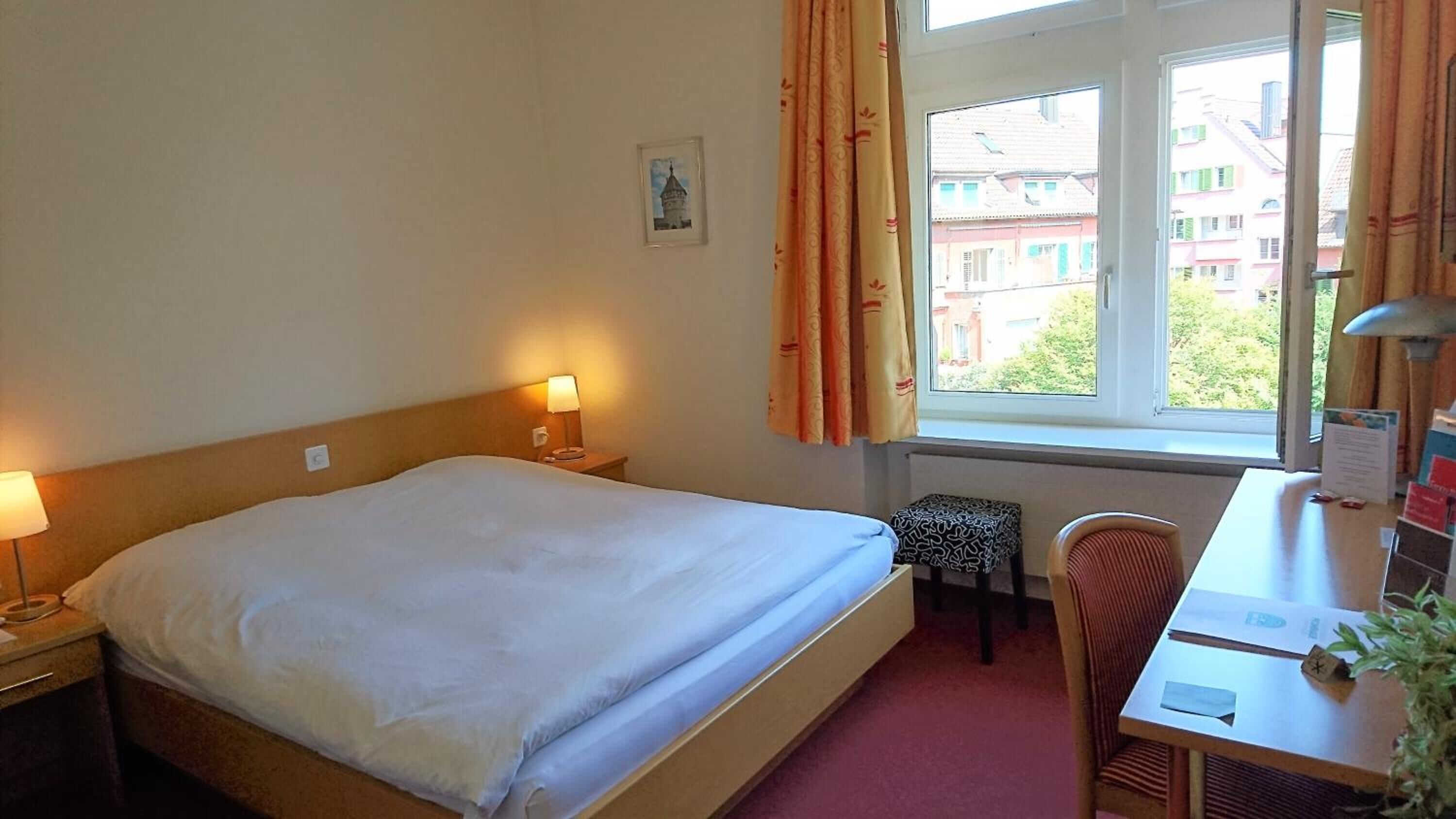 A hotel room with a bed, bedside lamps, a desk with a chair, a small table, and a window with curtains.