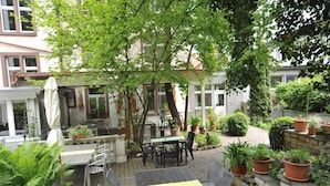 Un patio al aire libre con mesas y sillas, rodeado de plantas en macetas y una estructura de pérgola.
