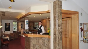Zona de recepción de un hotel con mostrador de madera, una recepcionista, comedor con mesas y sillas, y un pilar de piedra.