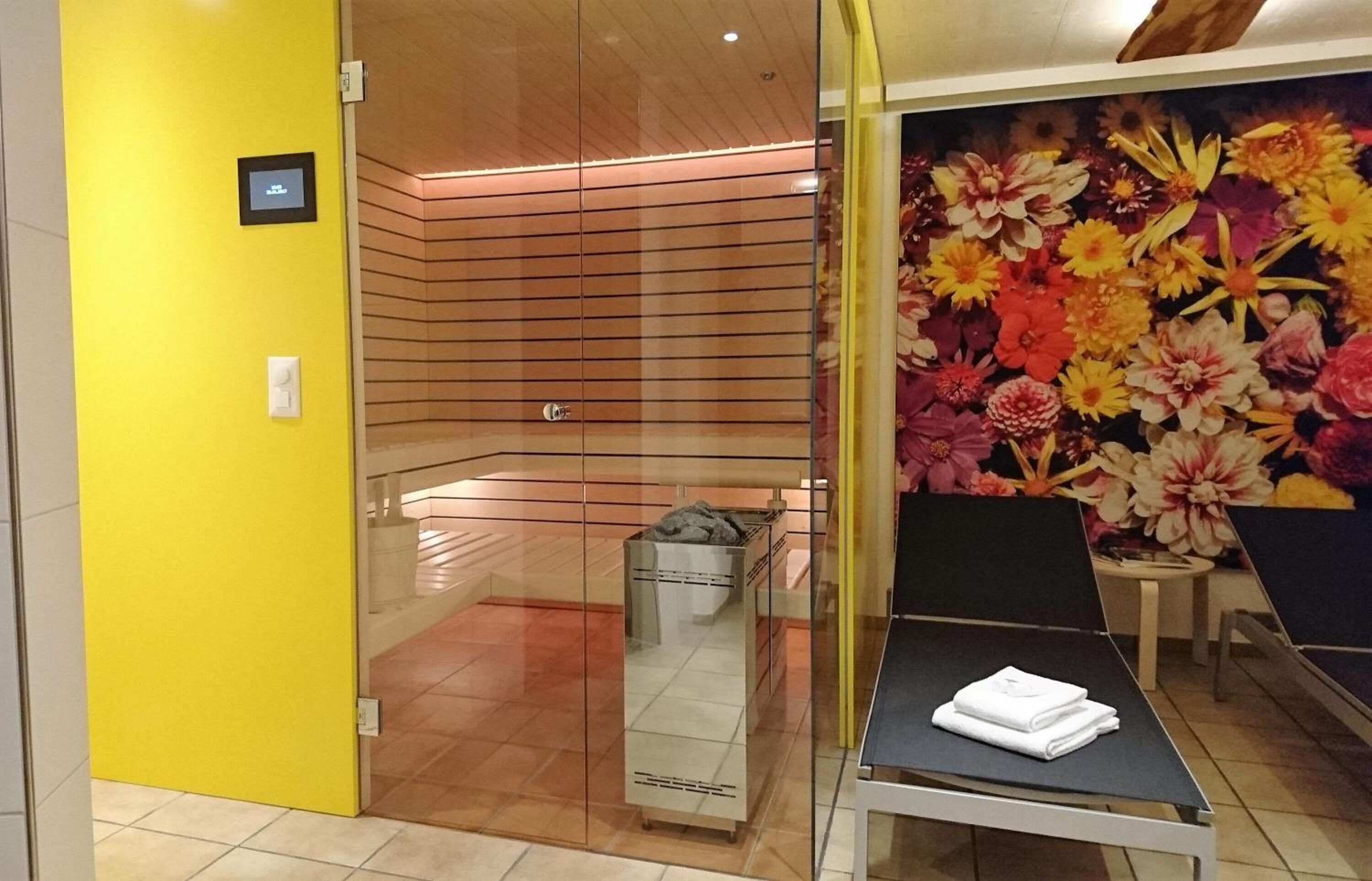 A room with a glass partition, a bed with a white towel, a floral wall mural, and a wooden ceiling.