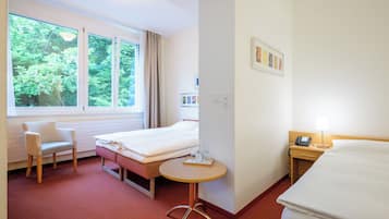 Et hotelværelse med seng, stol, lille bord, skrivebord med lampe og vindue med udsigt til træer.