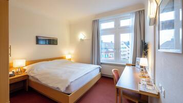 Een hotelkamer met een bed, nachtkastje, lamp, bureau, stoel en een raam met uitzicht op gebouwen.