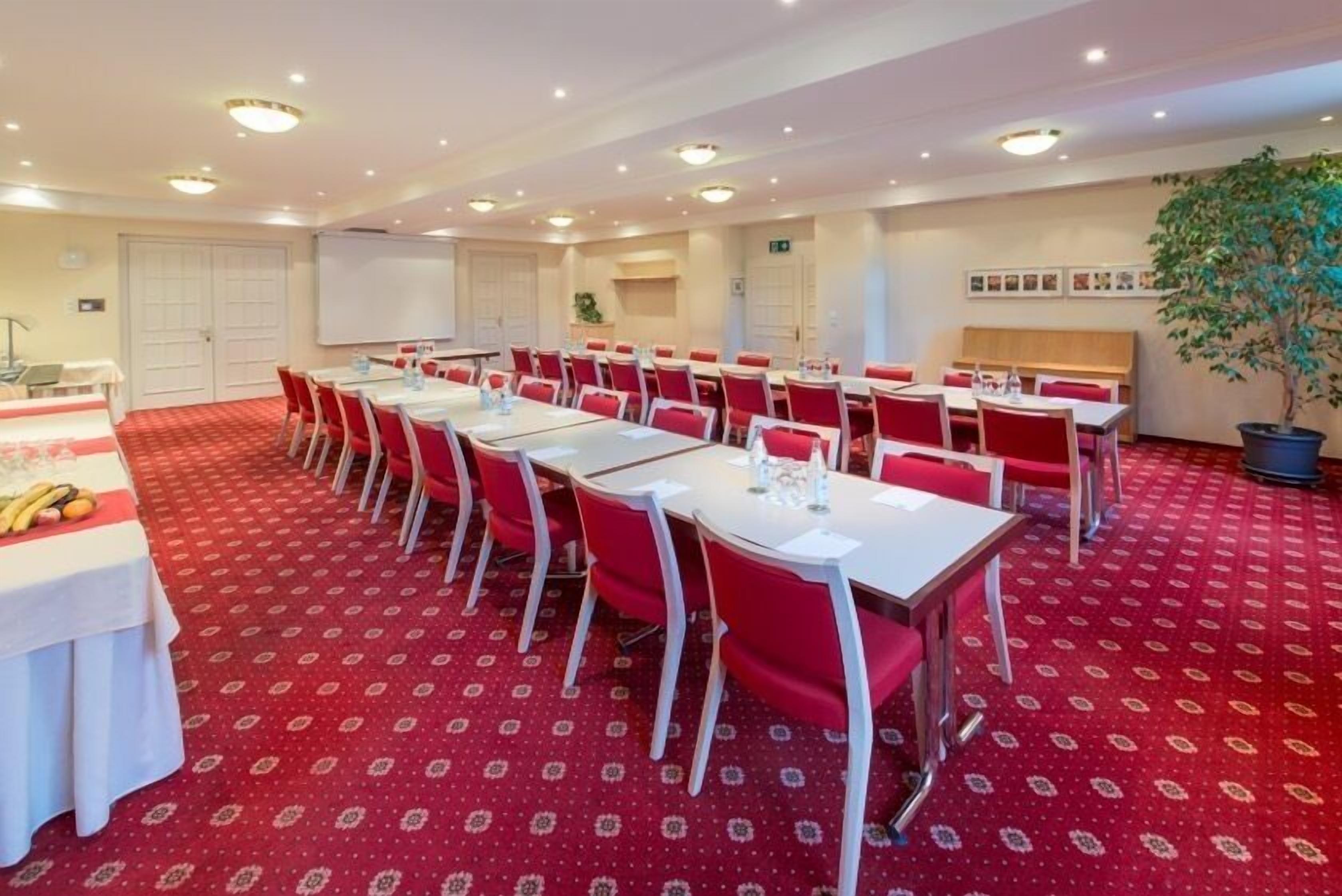 Una sala de conferencias con piso cubierto de alfombra roja, mesas blancas y sillas rojas.