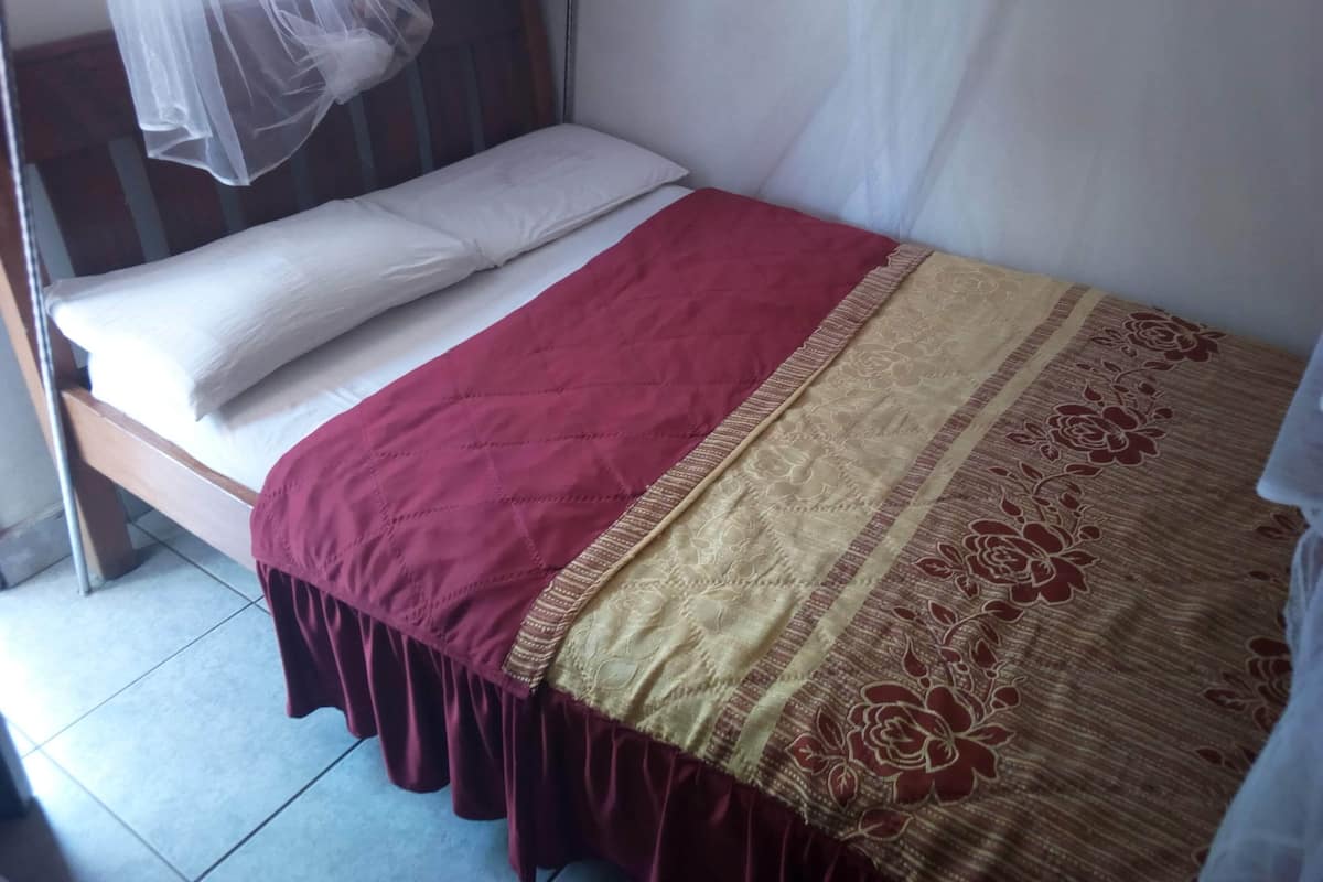 Un lit simple avec un matelas blanc et deux oreillers, une couvre-lit rouge et beige, et une moustiquaire au-dessus.