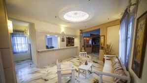 A spacious living area with a marble floor, a white sofa, a small table, and a bar area with shelves and a counter. - Hotel Saint George Valsamitis (Amorgos)
