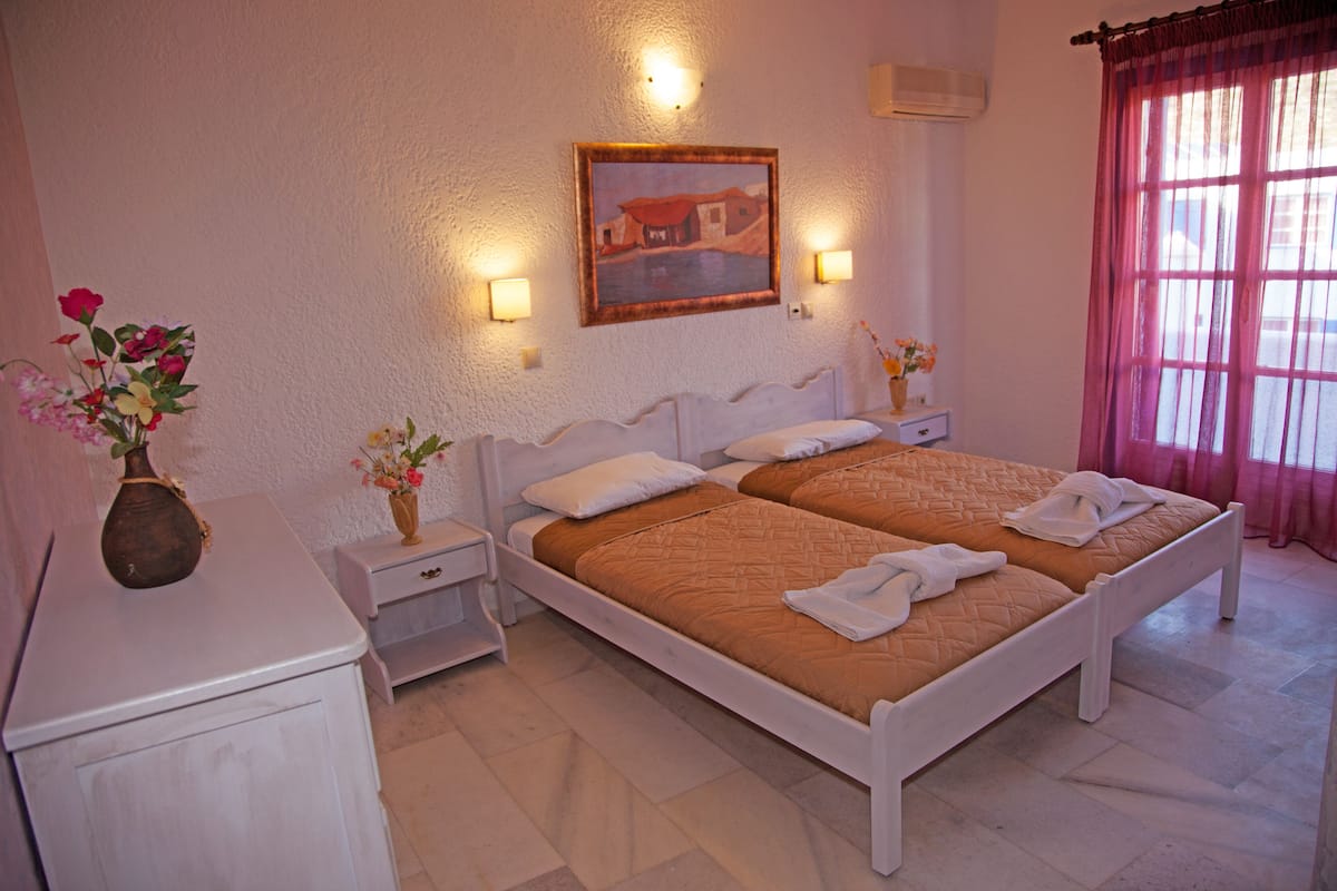 A bedroom with two beds, bedside tables, a nightstand, a painting on the wall, and a window with curtains.