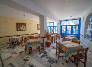 A dining area with multiple tables and chairs, a large vase with dried plants, and blue-framed windows. - Hotel Saint George Valsamitis (Amorgos)