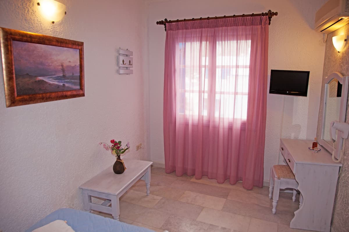 A room with a bed, a small table with a vase, a vanity with a mirror, and a window with pink curtains.