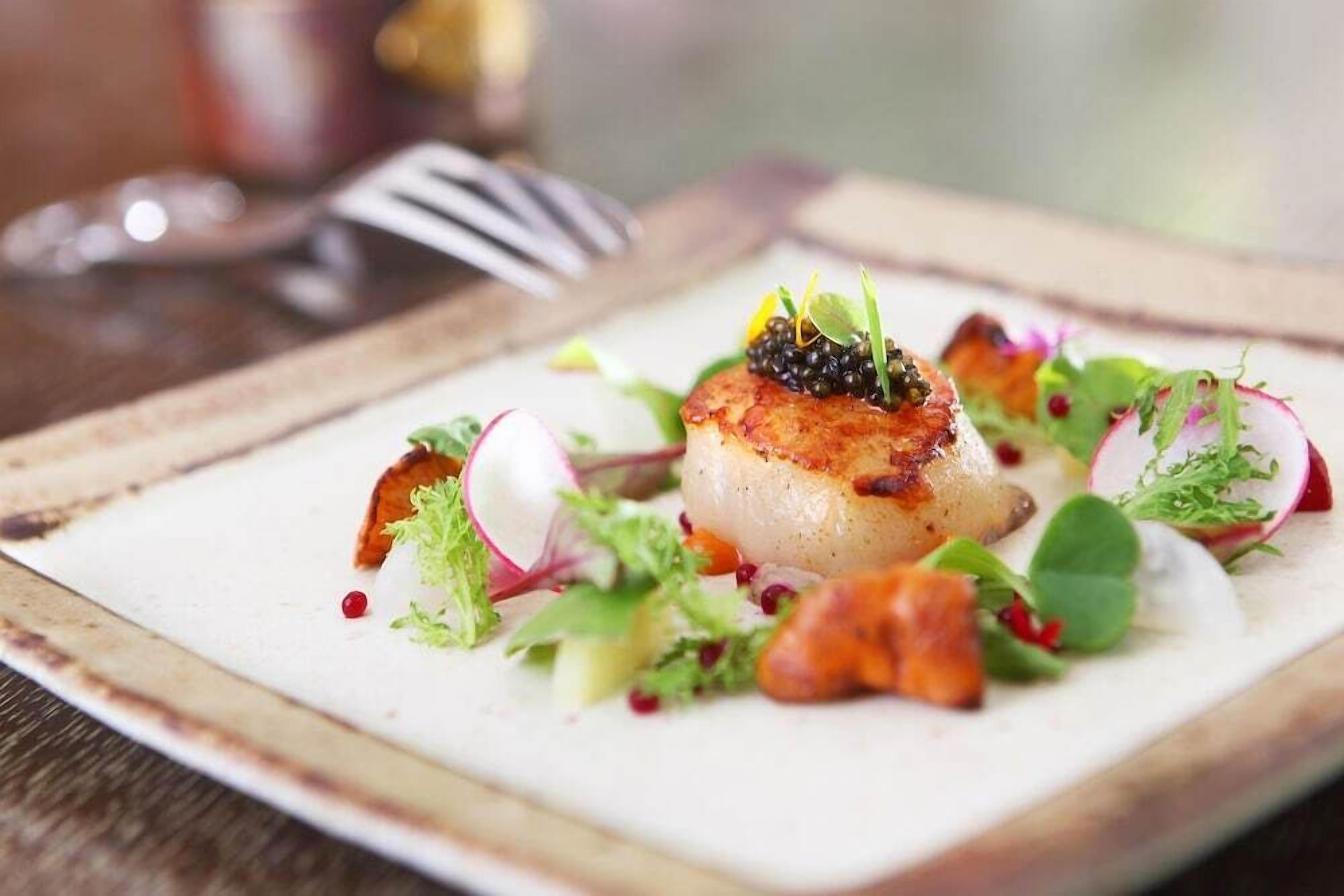 Un plato emplatado con vieira sellada, adornado con microgreens y caviar, servido sobre un plato blanco con un fondo de mesa de madera rústica.
