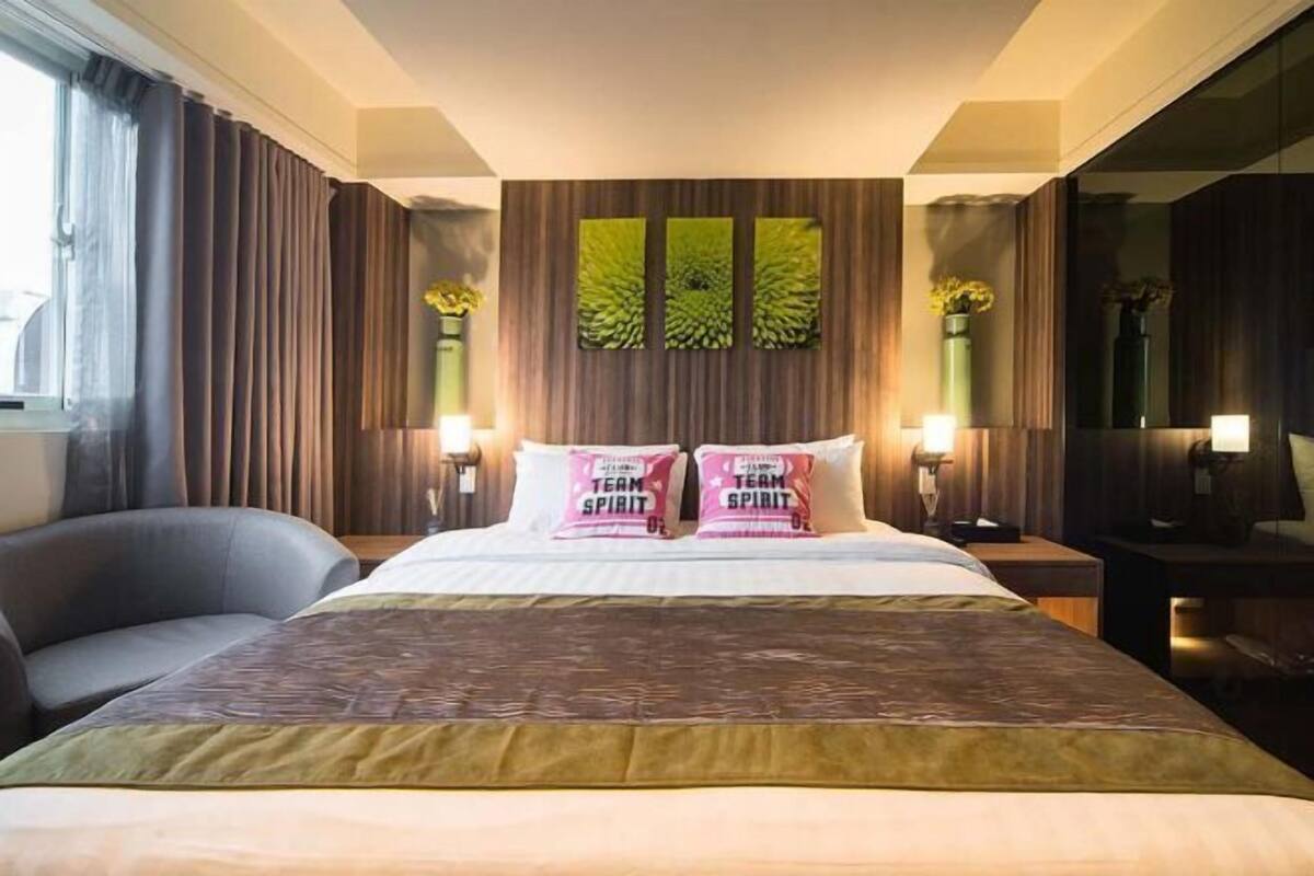 A hotel room with a large bed, two pillows with 'TEAM SPIRIT' text, a bedside table with a lamp, a chair, and a large mirror.