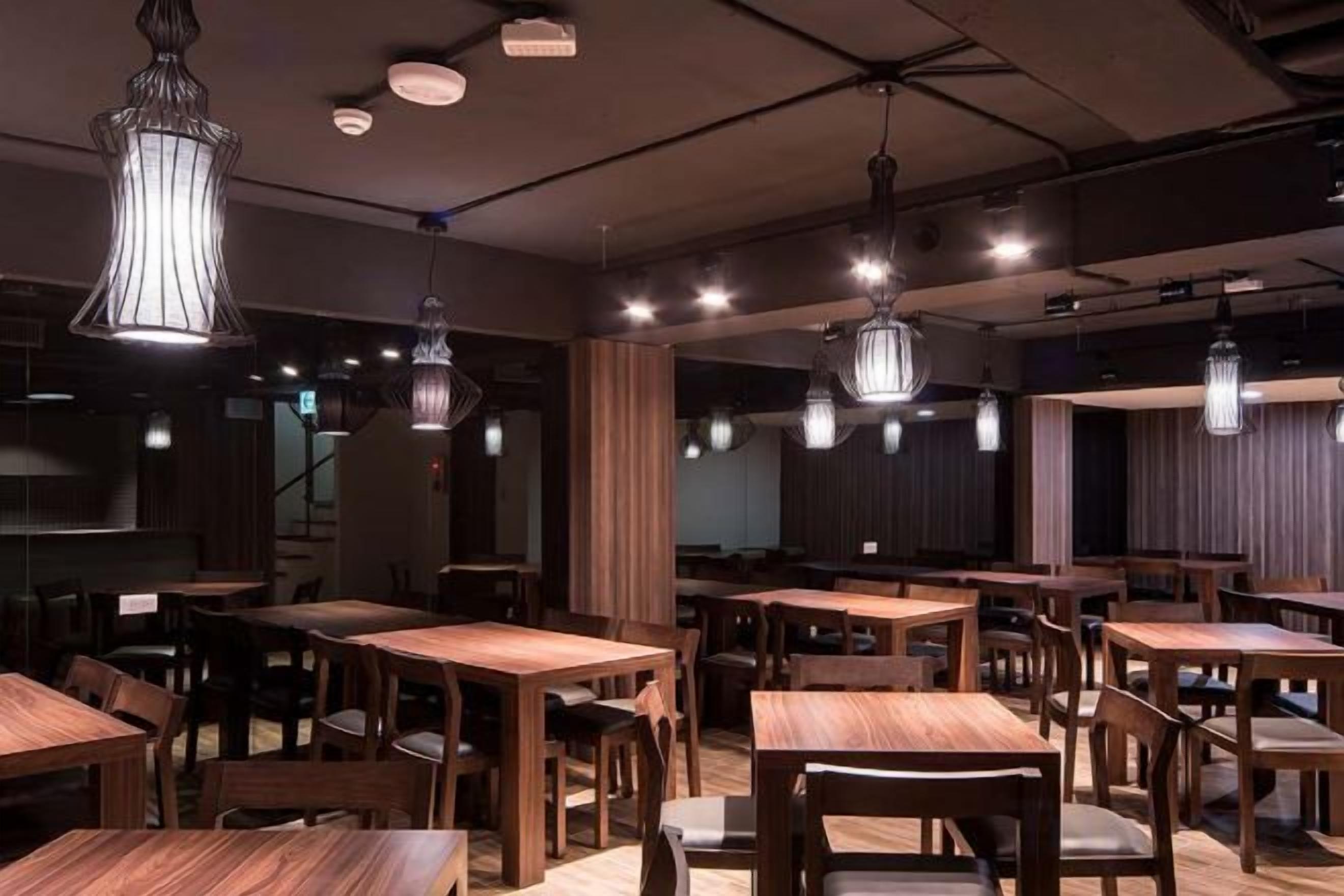 Interior de restaurante con varias mesas y sillas de madera, lámparas colgantes y una paleta de colores oscuros.