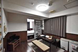 A compact hotel room with a bed, a desk, a bench, a small table, a television, and a refrigerator.