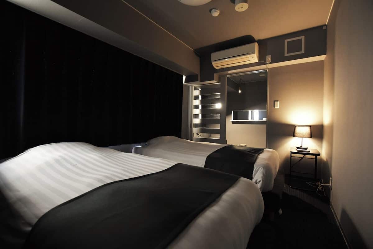 A hotel room with two beds, a wall-mounted air conditioner, a bedside table with a lamp, and a mirror.