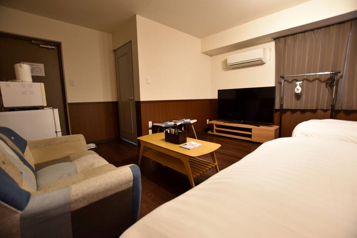 A hotel room with a bed, a TV, a desk, a chair, a refrigerator, and an air conditioning unit.