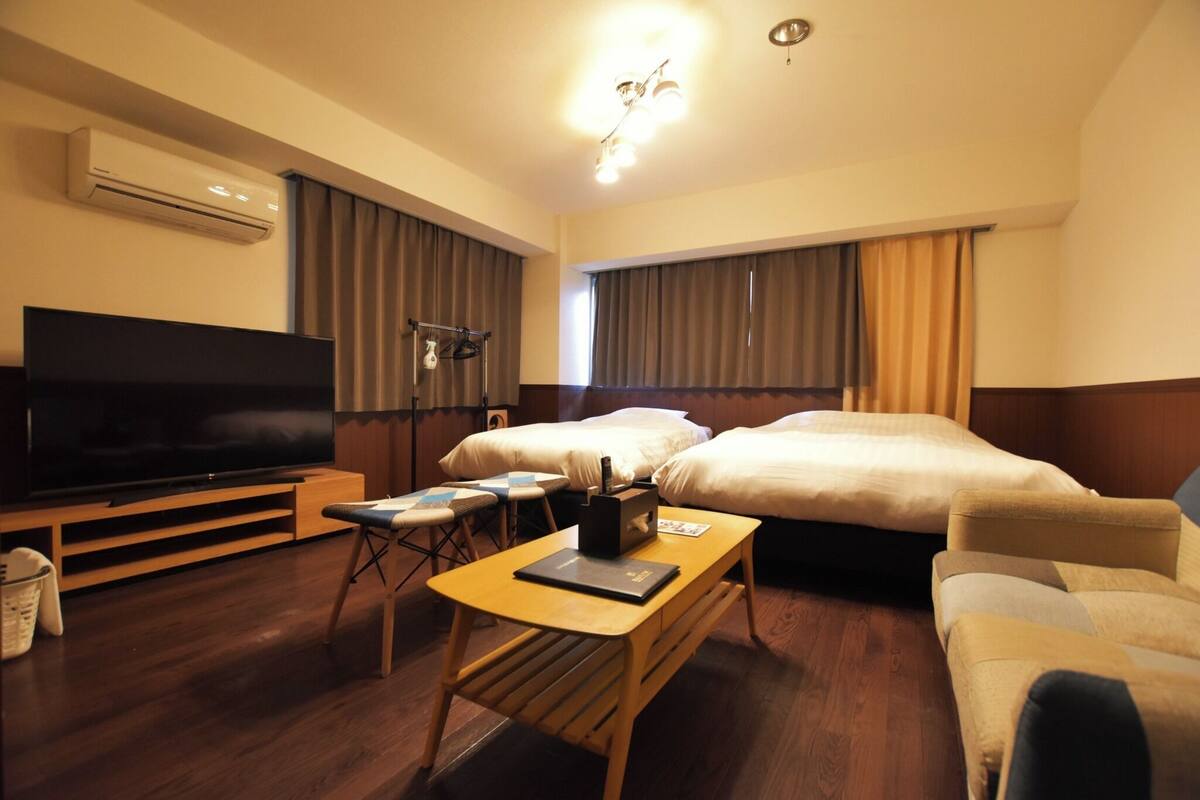 A hotel room with a flat-screen TV, a wooden table, two beds, a sofa, and an air conditioning unit.