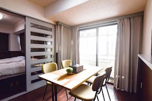 A hotel room with a bed, a desk, chairs, a small plant, and a window with curtains.