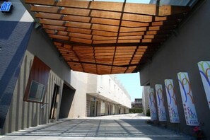 Une allée couverte avec un plafond en bois, menant à un bâtiment doté d’une façade moderne et d’une enseigne avec un logo.