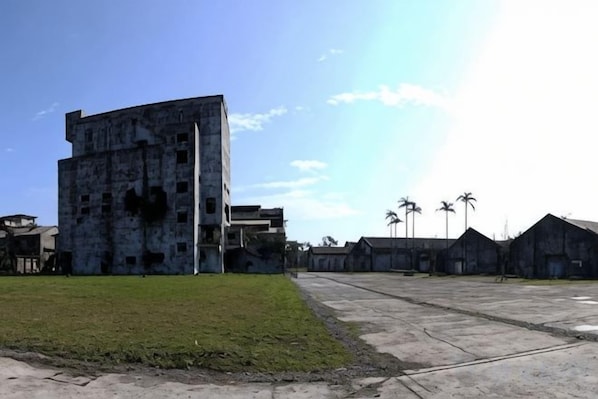 Un grand bâtiment à plusieurs étages, doté d’un toit plat, entouré d’une zone verdoyante et de constructions plus petites.