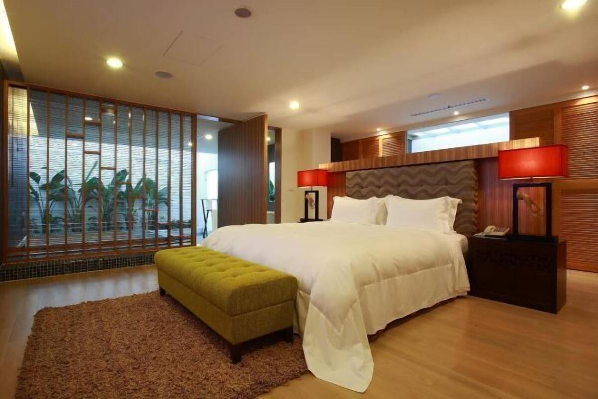 A modern bedroom with a large bed, a green ottoman, a wooden headboard, and a view through a shoji screen.