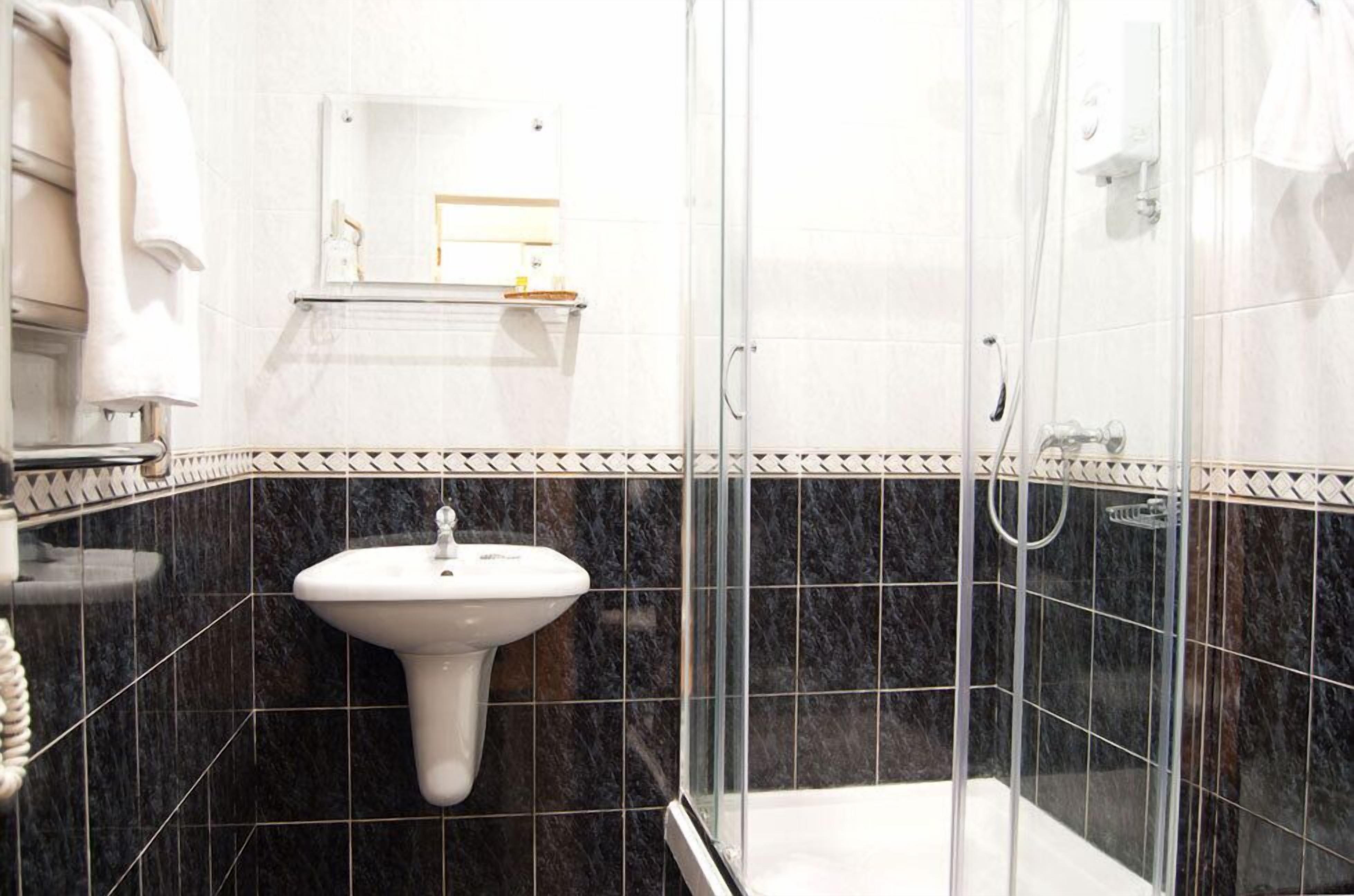 Un baño con ducha envuelta en vidrio, lavamanos pedestal blanco y suelo de baldosas.