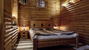 A log cabin bedroom with a wooden bed, a nightstand with a lamp, and a window with a view of trees. - Sonya Hotel (Kyiv)