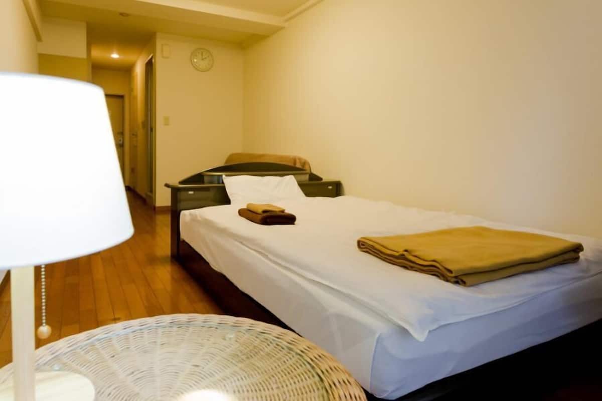 A small, neatly arranged bedroom with a single bed, a bedside table, a lamp, and a wicker chair.