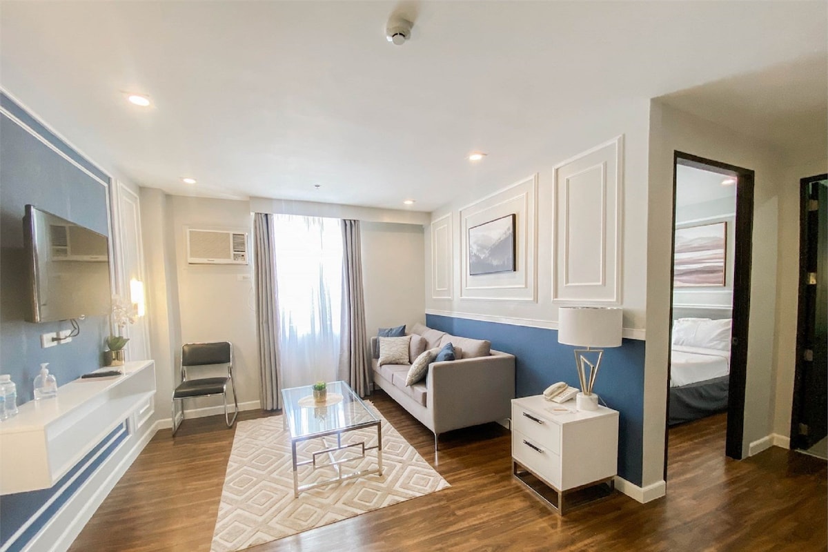 A modern hotel room with a sofa, a coffee table, a TV, a bedside table with a lamp, and a bed.