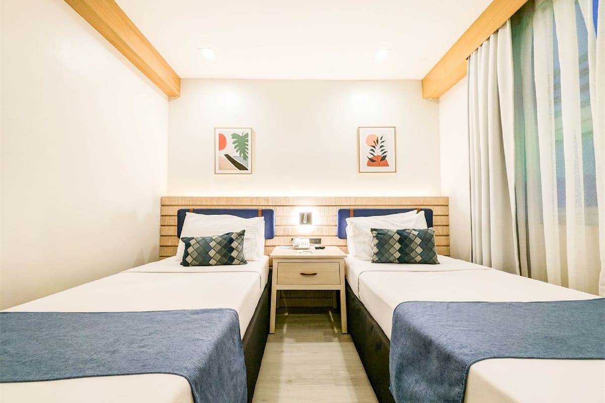 Une chambre d’hôtel avec deux lits simples, une table de chevet, un bureau et une fenêtre avec des rideaux.