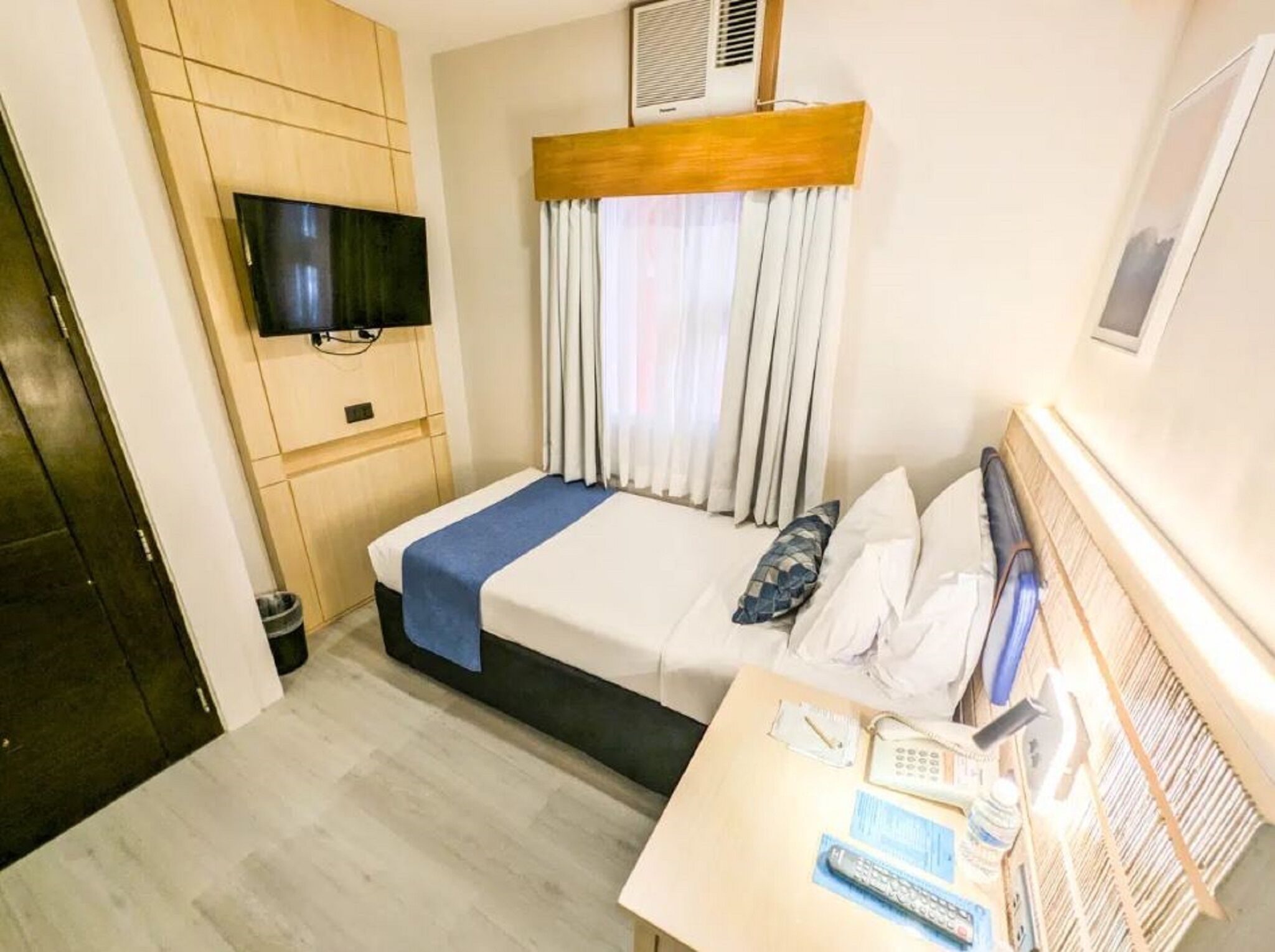 A hotel room with a bed, a TV mounted on the wall, a desk with a phone and remote control, and a window with curtains.