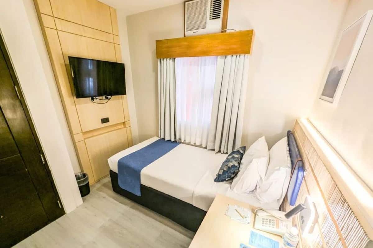 Une chambre d’hôtel comprenant un lit, un téléviseur fixé au mur, un bureau avec un téléphone et une télécommande, ainsi qu’une fenêtre avec des rideaux.