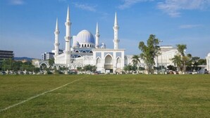 Une grande mosquée dotée de plusieurs minarets et d’un dôme, entourée d’un champ verdoyant et de palmiers.