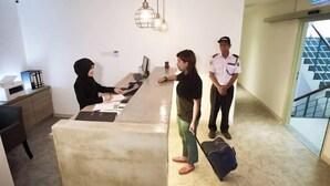 Une réception avec une réceptionniste, un visiteur et un agent de sécurité. On y voit un bureau avec un ordinateur et des documents, un luminaire suspendu et un escalier en arrière-plan.