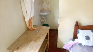 Ein schlichter, kleiner Raum mit Holztisch, Bett mit gemustertem Kopfkissen, Wandwaschbecken und Vorhang.