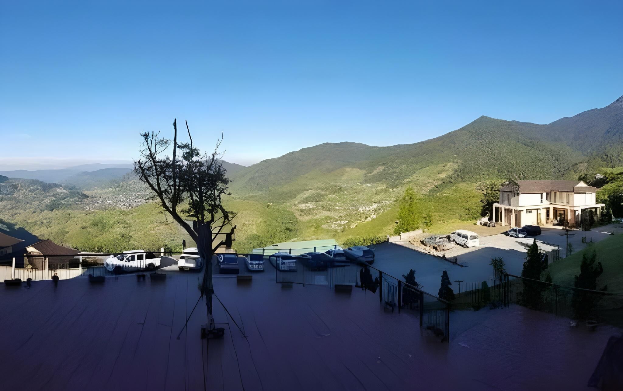 A parking area with several cars, a tree, and a building with a view of green hills in the background.