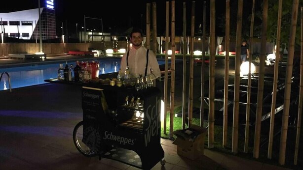 Un espace bar extérieur, aménagé pour la nuit, avec un chariot portant l’inscription « Schweppes » et une piscine en arrière-plan.
