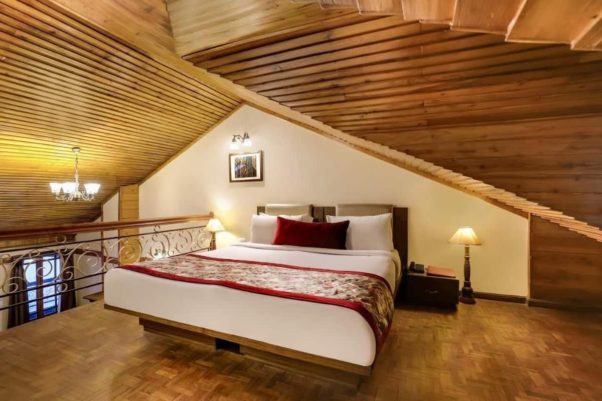 A bedroom with a sloped ceiling, wooden beams, a bed with a red pillow, two bedside lamps, and a framed picture on the wall.