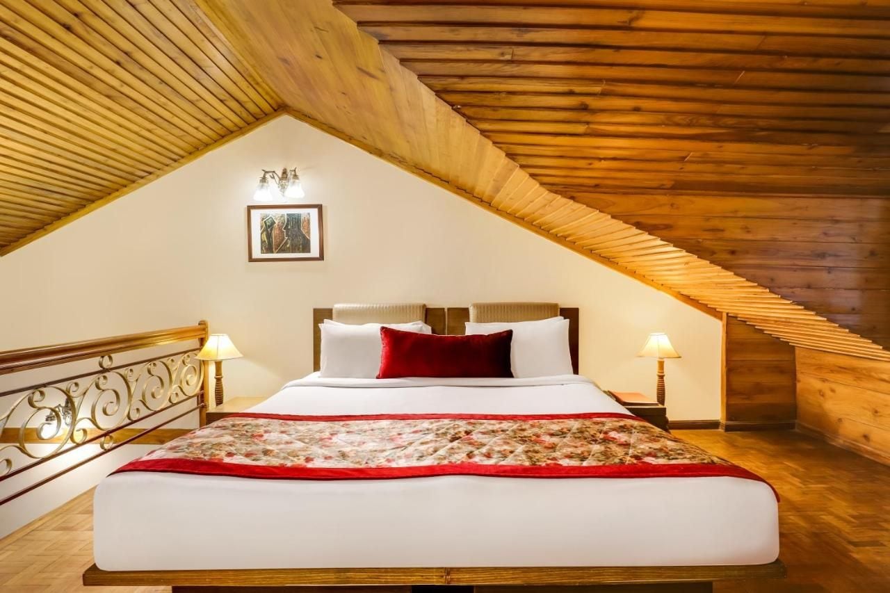 A bedroom with a sloped ceiling, a bed with white linens and a red throw, two bedside lamps, and a framed picture on the wall.