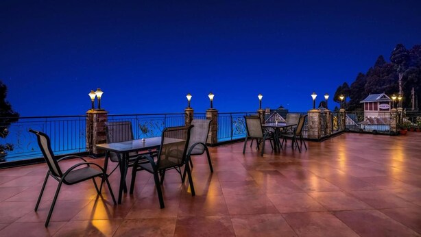 An outdoor dining area with tables and chairs, a railing, and a view of the water. - Summit Grace Hotel Spa (Darjeeling)