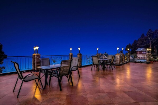 An outdoor dining area with tables and chairs, a railing, and a view of the water. - Summit Grace Hotel Spa (Darjeeling)