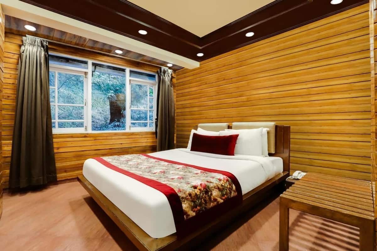 A room with a wooden paneled wall, a large bed with a floral bedspread, a bedside table, and a window with curtains.