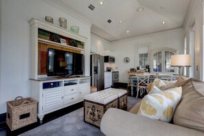 Uma sala de estar moderna com televisão, armários brancos, uma área de jantar e uma cozinha ao fundo.