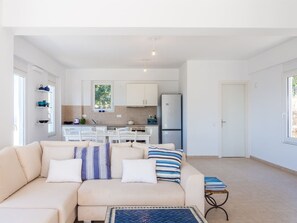 Un salon moderne avec un canapé beige, une petite table basse et une vue sur un coin cuisine doté de meubles blancs et d’un réfrigérateur.
